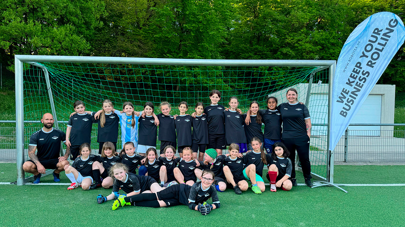 Die U11-Mädchenmannschaft posiert mit ihrem Trainerteam in einem Fußballtor. Rechts von ihnen steht eine Beachflag mit Bobotex-Werbung.